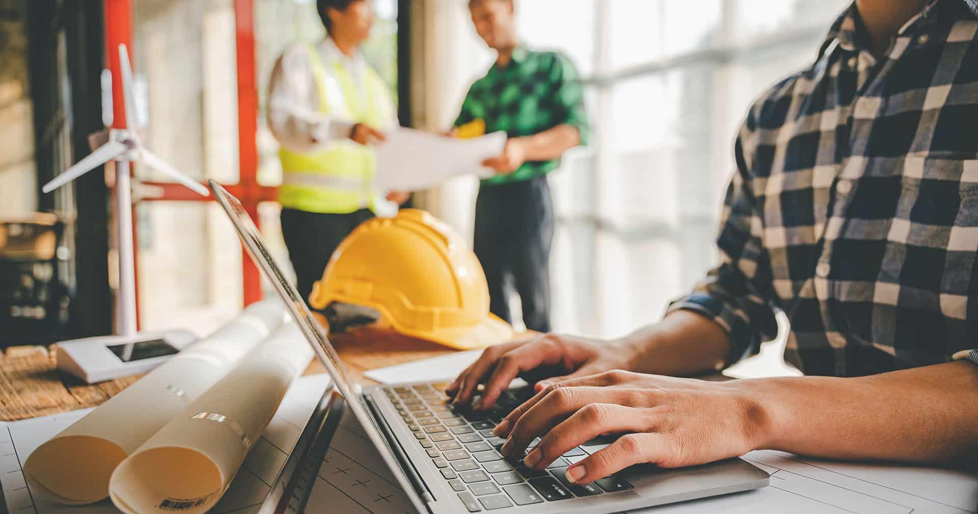 Mâinile unui meseriaș tastând pe un laptop, lângă o cască galbenă și planuri rulate, cu doi colegi discutând peste o schiță în fundal