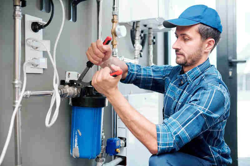 Instalator în cămașă în carouri și șapcă albastră strânge cu cleștele racordul unui filtru de apă montat pe o instalație de țevi metalice