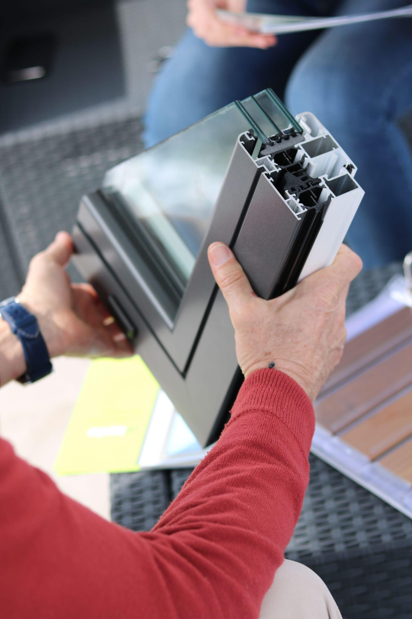 Mâini care țin o secțiune de profil de fereastră din aluminiu cu geam triplu, cu camerele interioare ale profilului vizibile, prezentată unui client