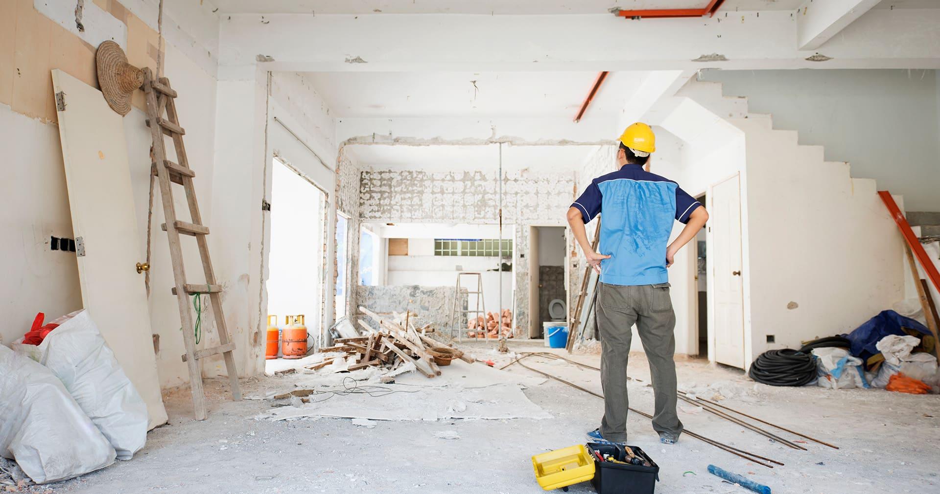 Muncitor cu cască galbenă, văzut din spate cu mâinile în șolduri, privind un apartament demolat până la structură, cu moloz, scări de lemn și trusa de scule pe jos