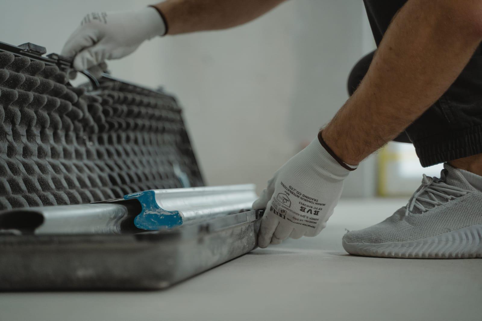 Mâini în mănuși albe care scot o riglă lungă de aluminiu dintr-o cutie de scule, pe o șapă gri într-o cameră la gri