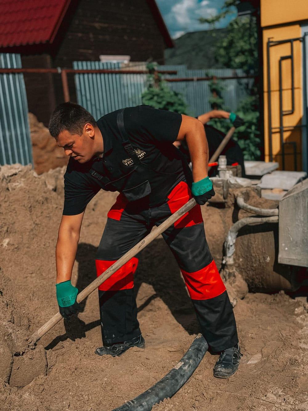 Muncitor în salopetă neagră cu roșu care nivelează pământul cu o unealtă cu coadă lungă, cu furtunul pompei de beton la picioare și un gard de tablă în fundal