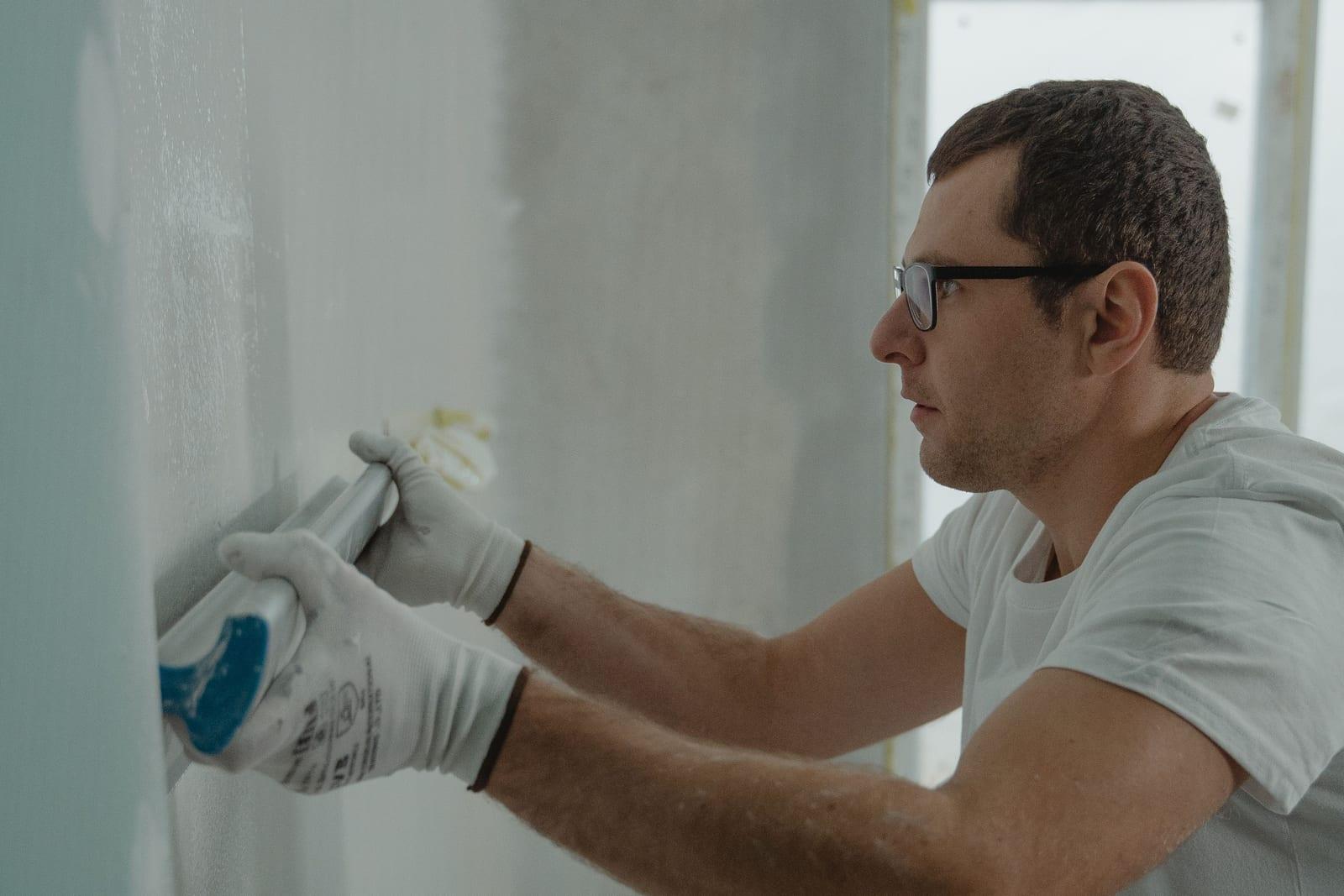 Meseriaș cu ochelari și mănuși albe care netezește cu un șpaclu lat cu mâner de aluminiu un perete interior gri