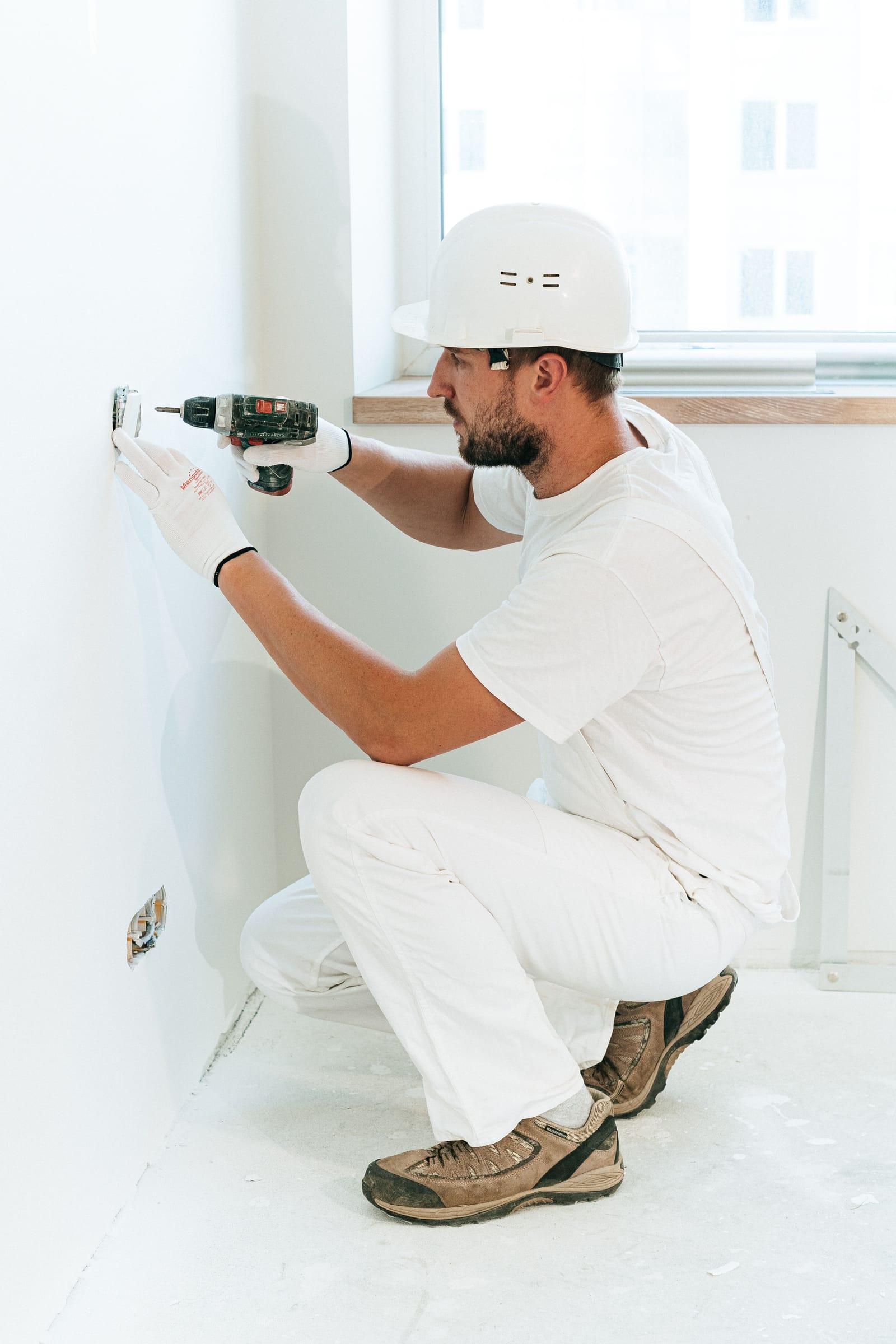 Electrician cu cască albă și mănuși, ghemuit lângă perete, montând cu bormașina o doză de priză într-o cameră proaspăt zugrăvită
