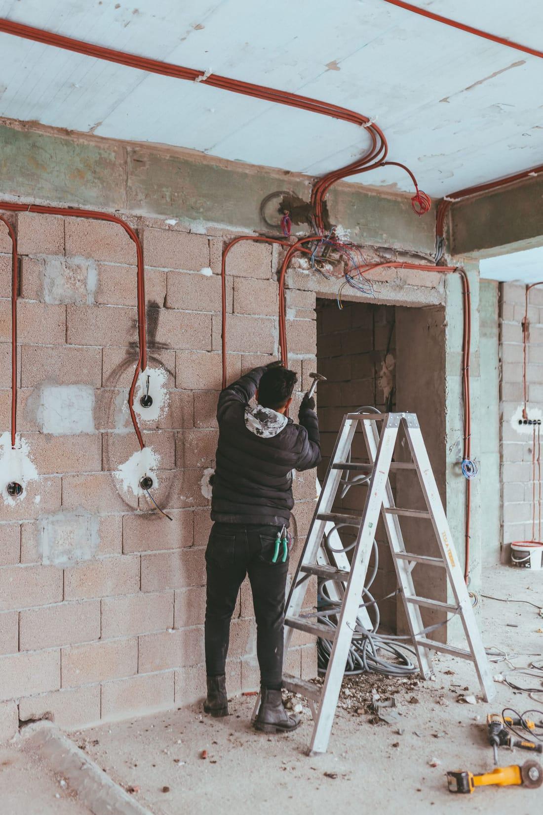 Electrician lucrând într-un apartament la roșu, cu trasee de tub copex portocaliu îngropate în zidărie și în tavan, doze montate în perete și o scară dublă alături