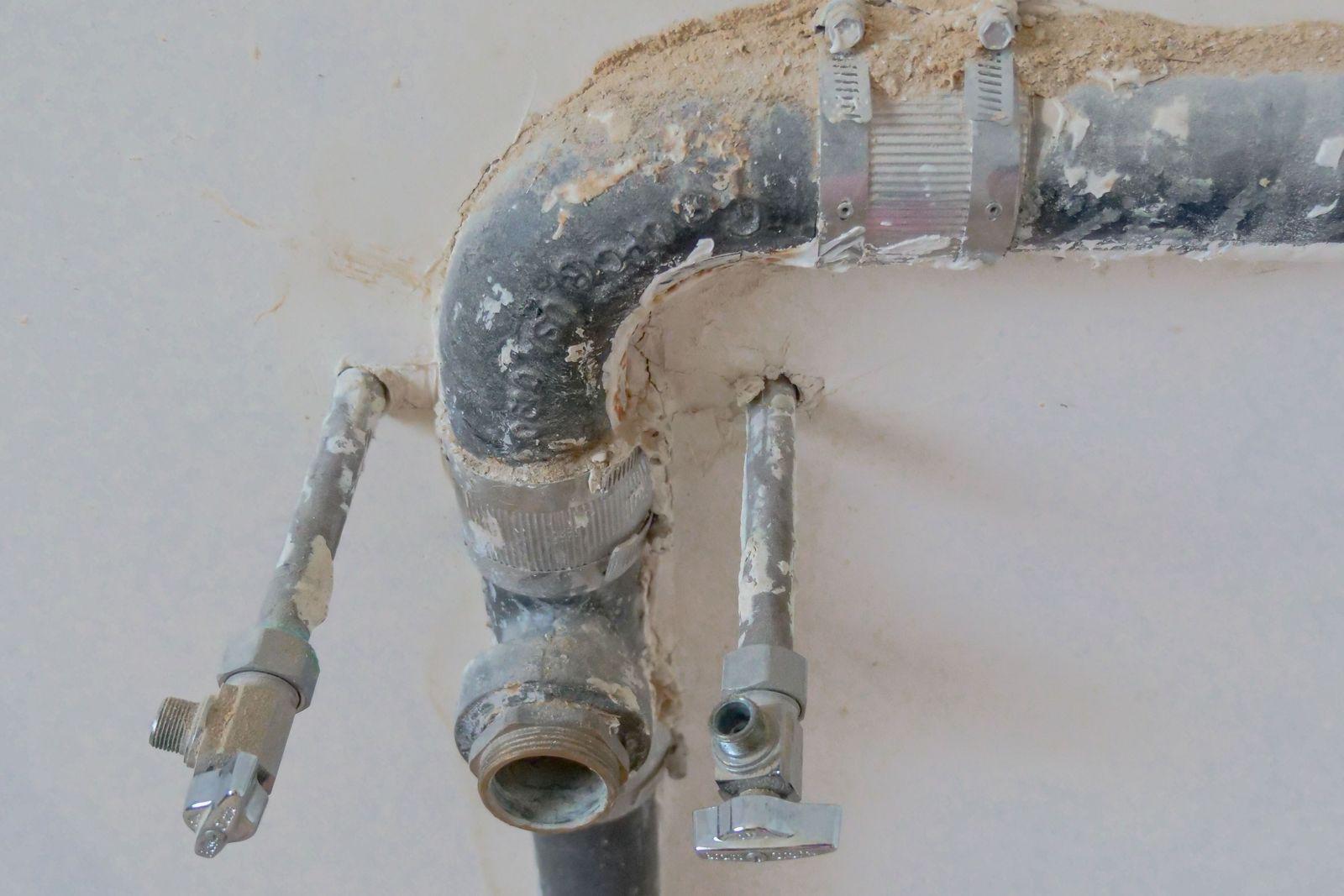 Instalație veche de fier zincat scoasă la iveală după decopertare, cu țeavă groasă prinsă în coliere, două robinete colțar cromate racordate pe lateral și un ștuț filetat lăsat liber, totul acoperit de praf de glet