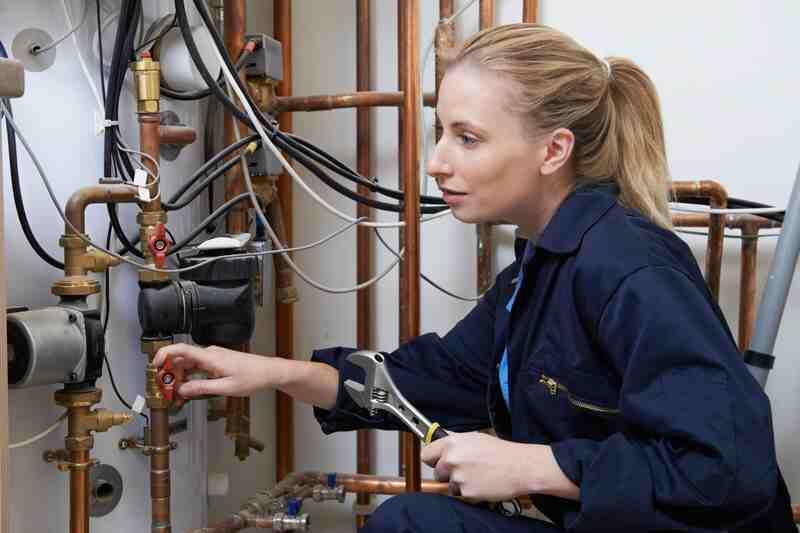 Instalatoare în salopetă bleumarin strânge cu o cheie reglabilă racordul unui boiler, pe o instalație de țevi de cupru cu robineți roșii