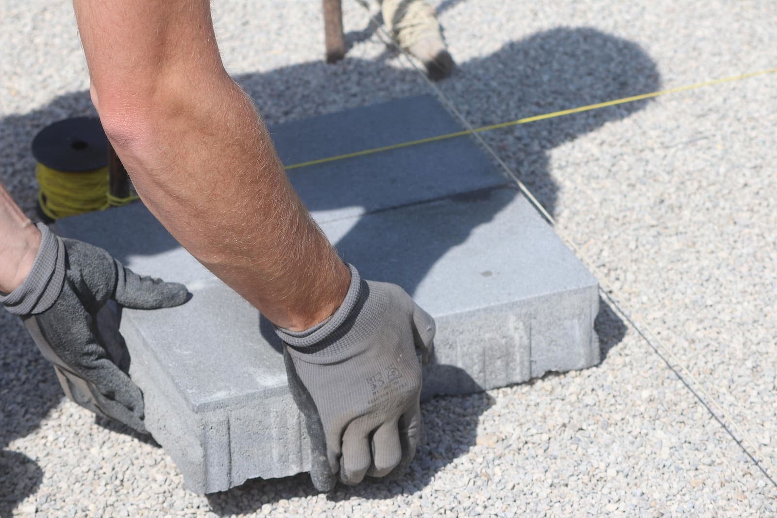 Muncitor cu mănuși de protecție care așază o pavelă de beton pe patul de split, cu sfoara de trasare întinsă alături