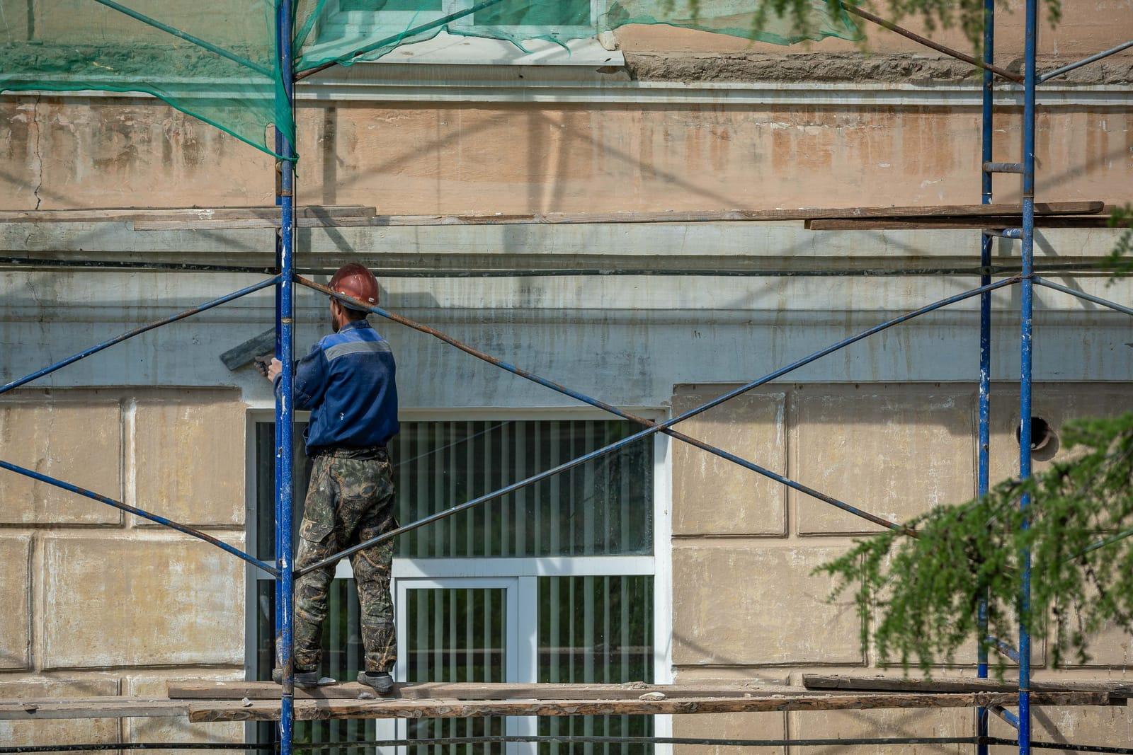 Muncitor cu cască roșie, pe o schelă metalică albastră, aplicând cu drișca un strat de tencuială pe fațada unei case, deasupra unei ferestre cu tâmplărie albă